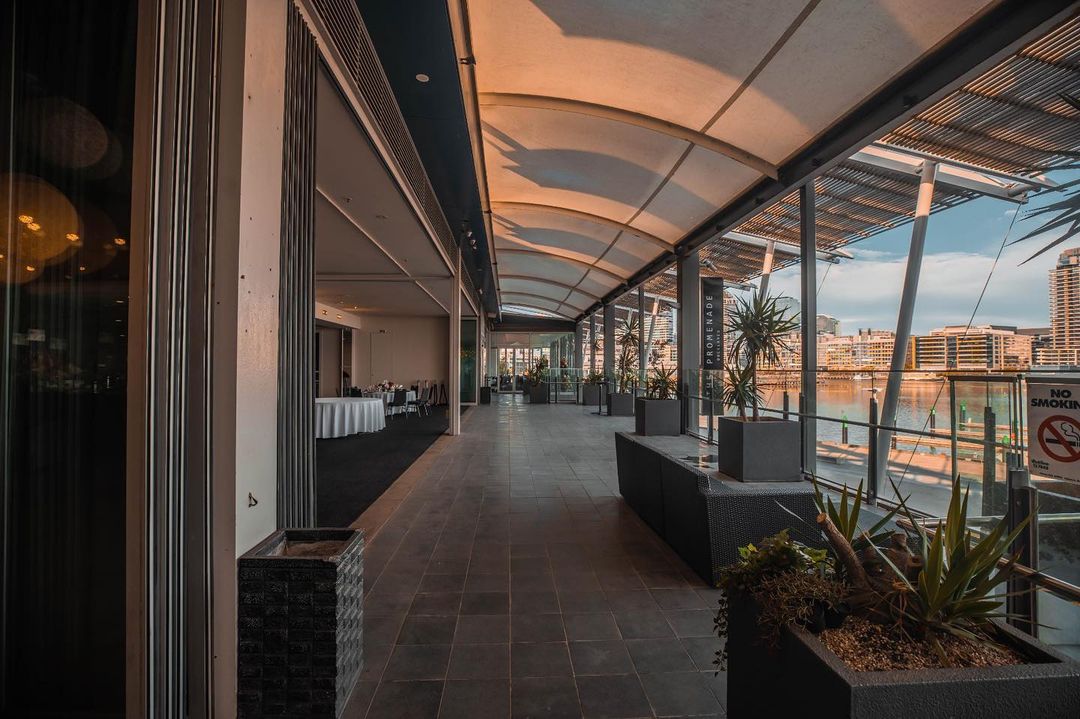 A scenic walkway with lush plants and tables in the middle at The Promenade Docklands Waterfront Room.