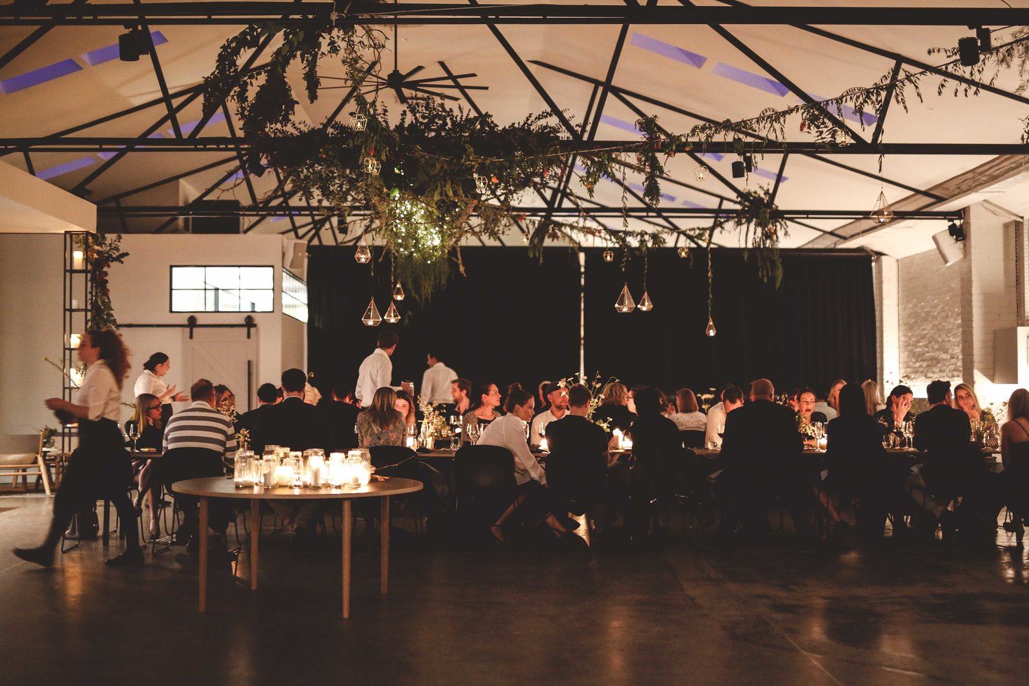 A lively scene at a dinner party venue in Melbourne, as a multitude of people gather around tables in a spacious room, enjoying each other's company.